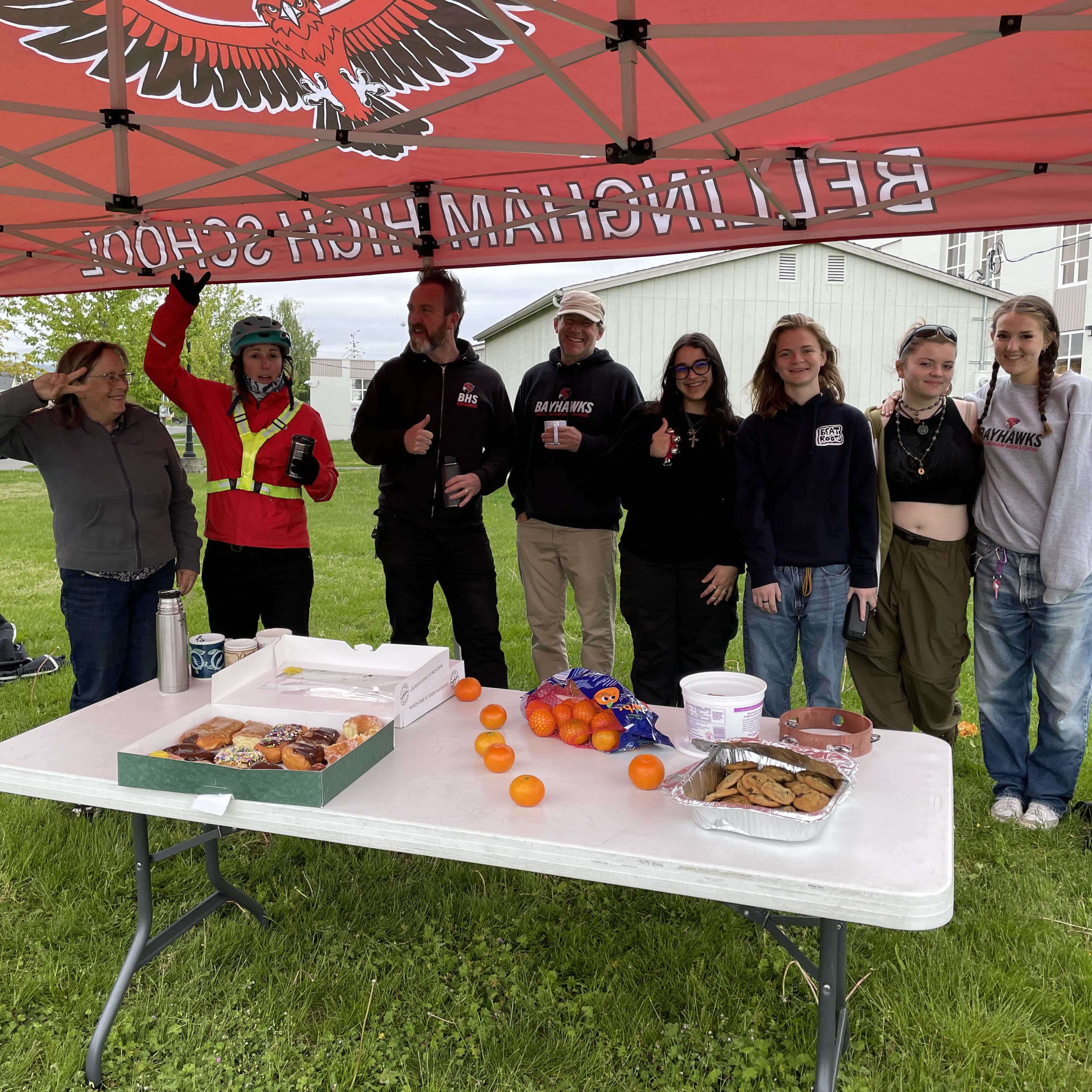 Several adults and high school students standing under a red tent behind a table with donuts and oranges